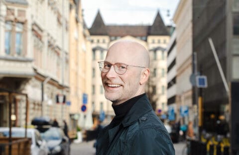 Kuvassa mies hymyilee etualalla ja taustalla näkyy kerrostaloja.